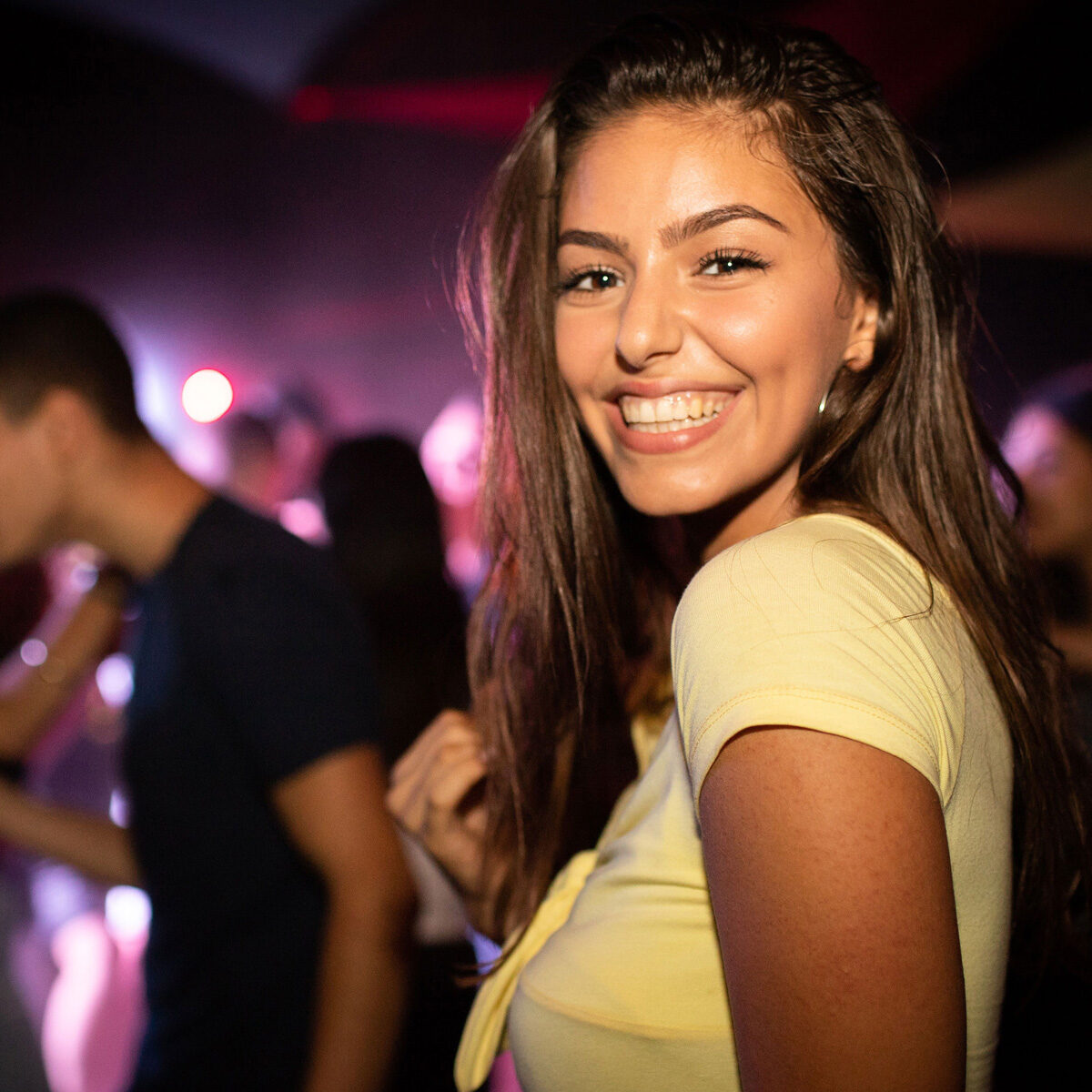 Una chica latina joven y de hermosa sonrisa posa de lado en una pista de baile, aparentemente en una discoteca. Tiene una camiseta pegada amarilla de mangas cortas y detrás se nota la dinámica social de un sitio nocturno.