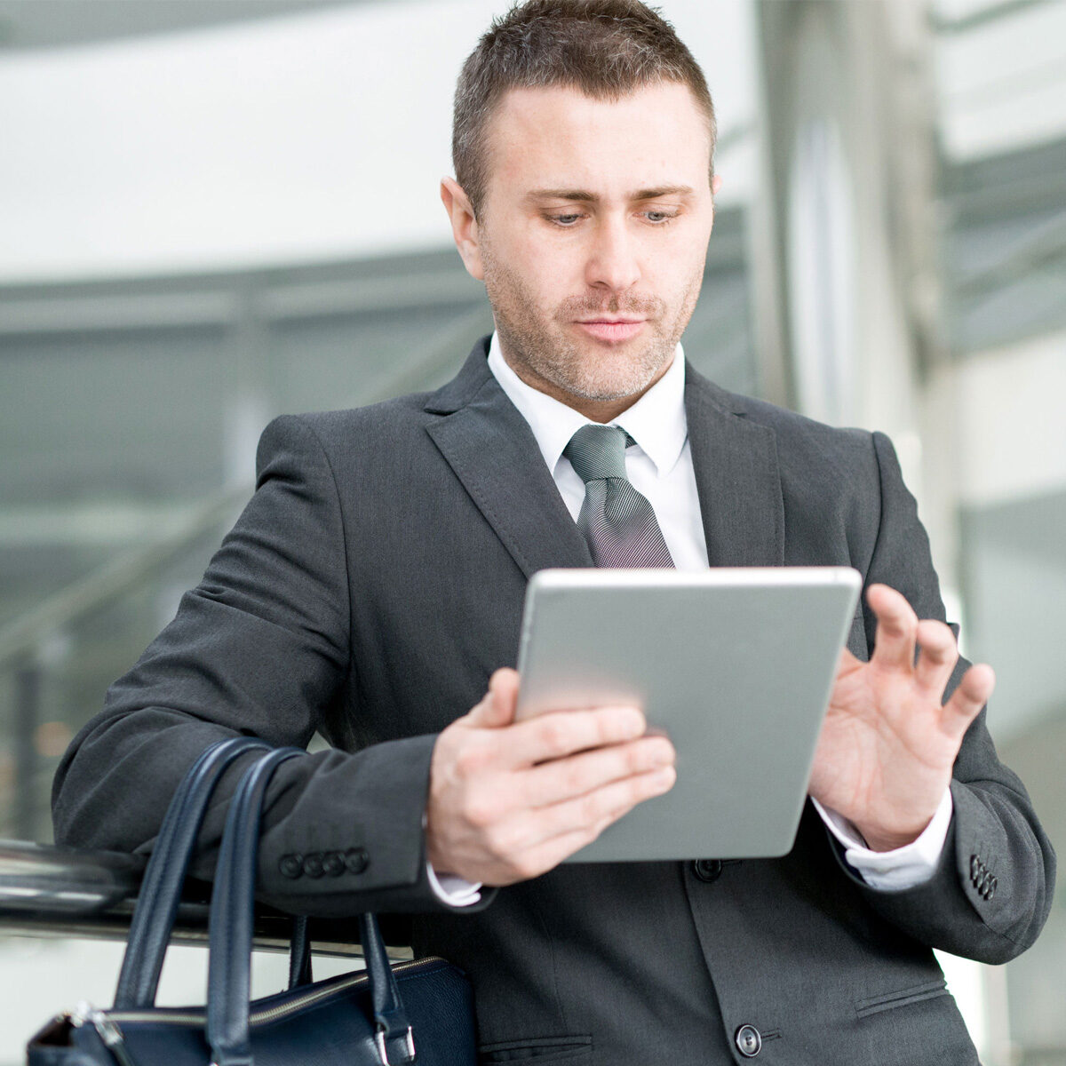 Un ejecutivo joven y de apariencia prolija revisa una tablet, usa un traje y tiene colgado de su brazo un maletin con asas de una cartera o bolso de cuero negro.