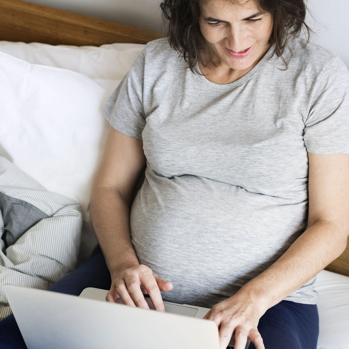 Una señora embarazada en sus treinta usa camisón gris, está sentada en una cama con almohadas blancas y un laptop en su regazo.