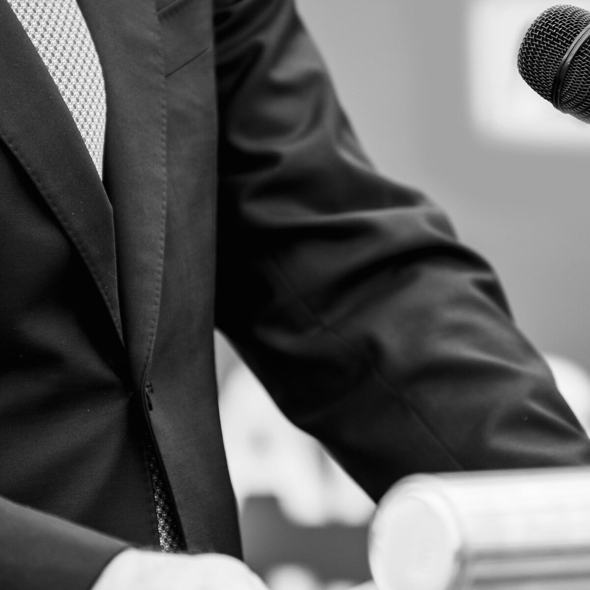 Se nota en closeup un traje de vestir masculino y un micrófono frente a este. Simula ser un orador en un podio, la foto está en blanco y negro.